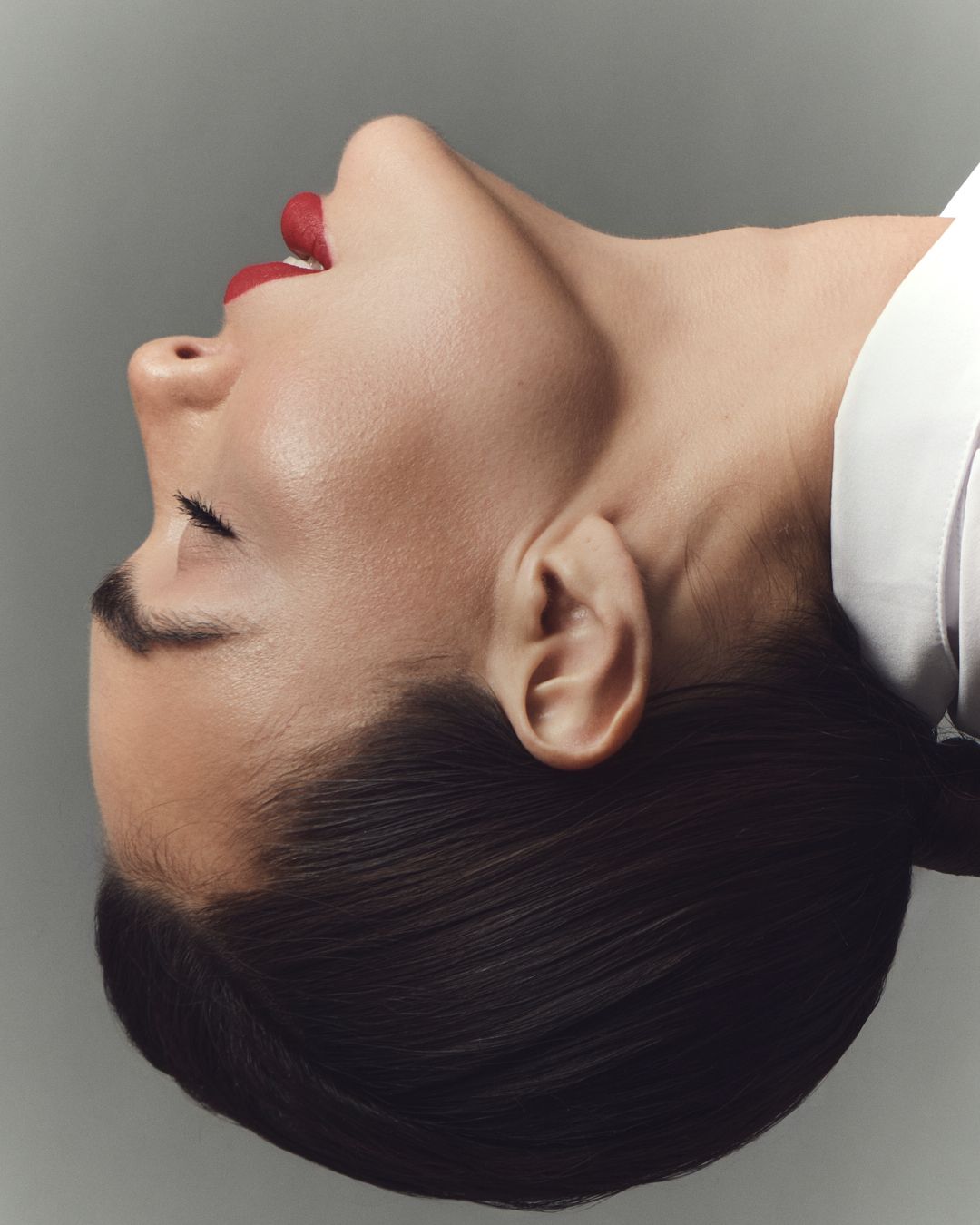 A woman with dark hair and red lips poses with her head tilted back, eyes closed, in a sleek editorial style.