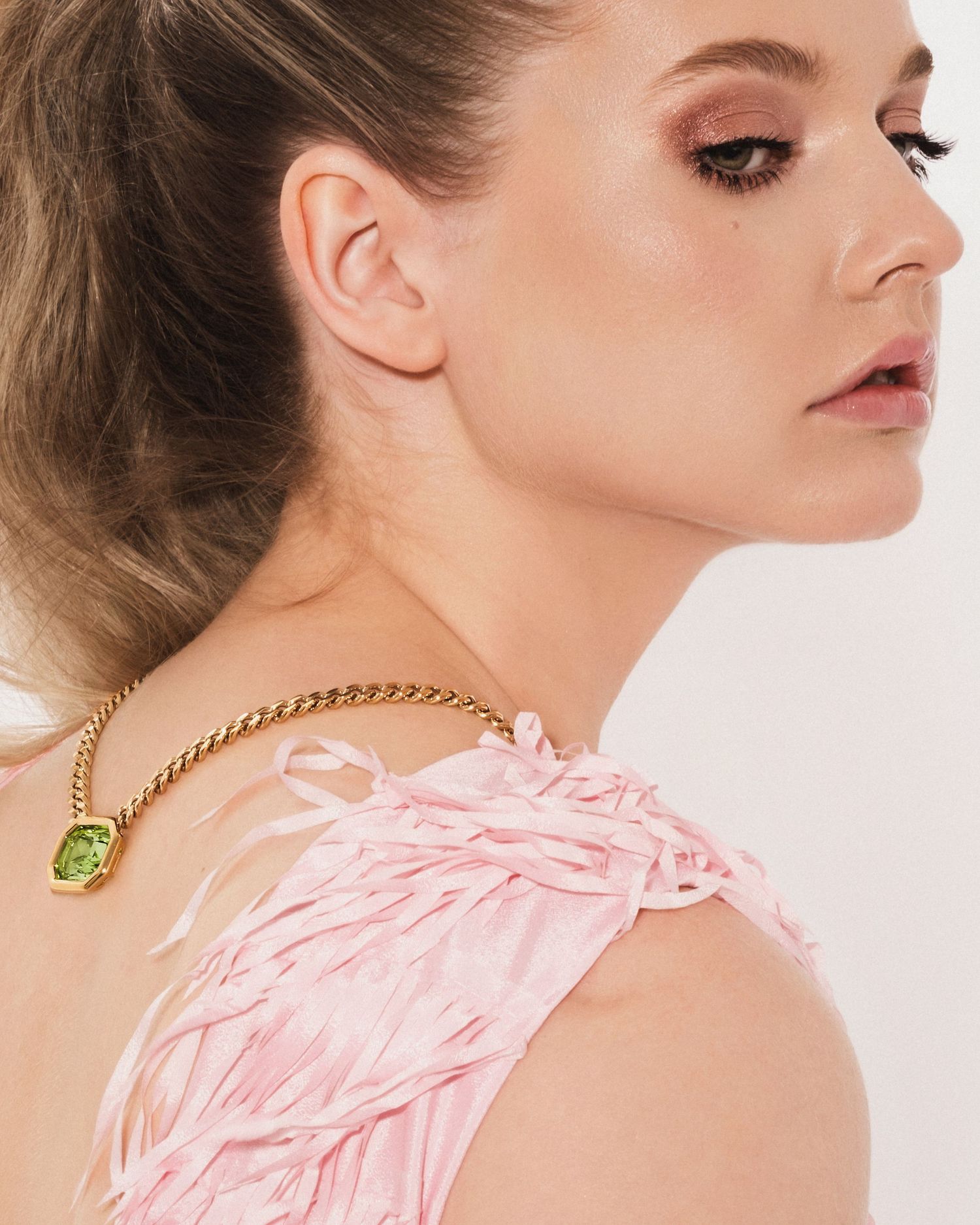 Woman in pink feathered dress with gold chain necklace featuring green gemstone pendant, smoky eye makeup.