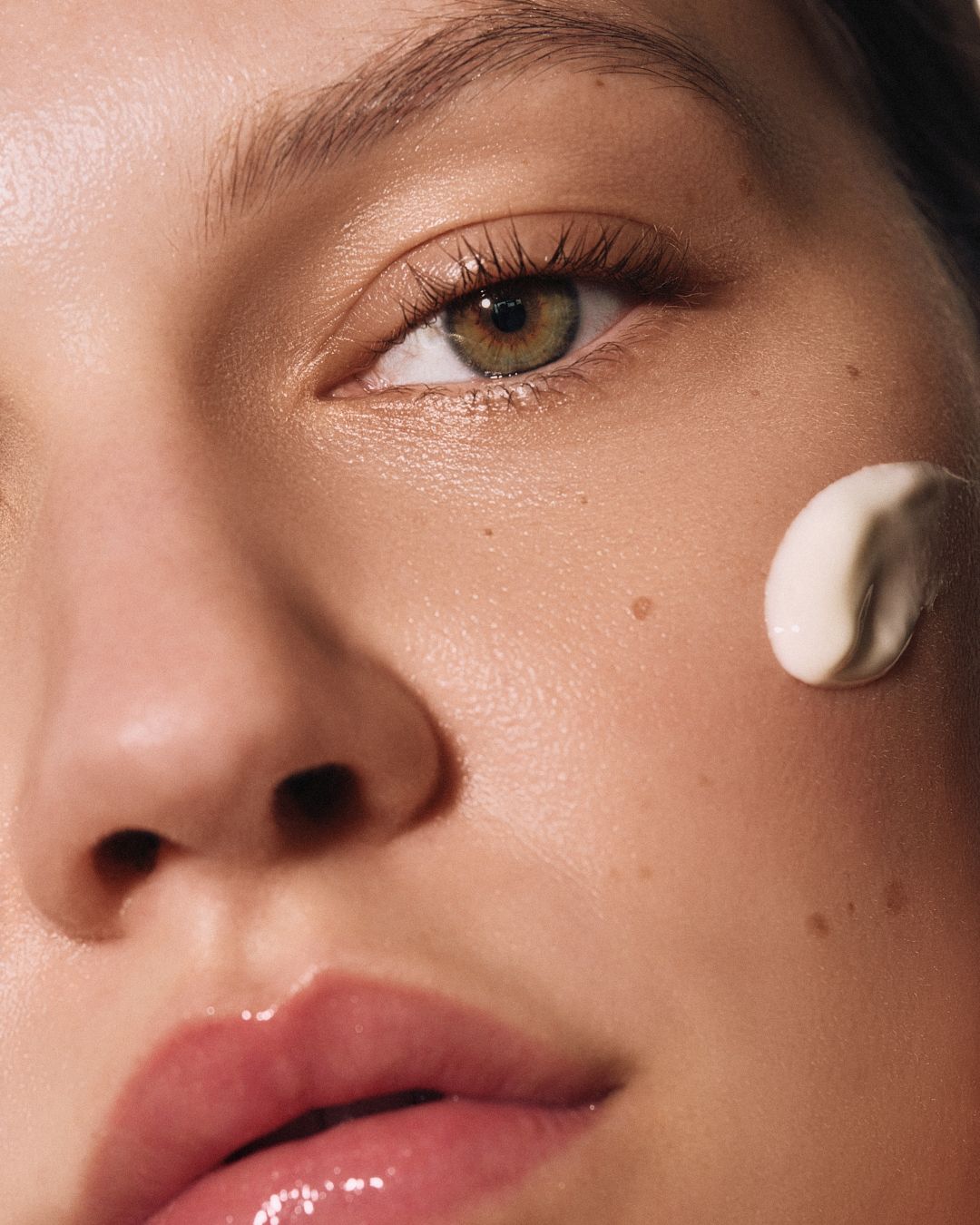 Extreme close-up of a woman's green eye and cheek with a dollop of white moisturizing cream applied to skin.