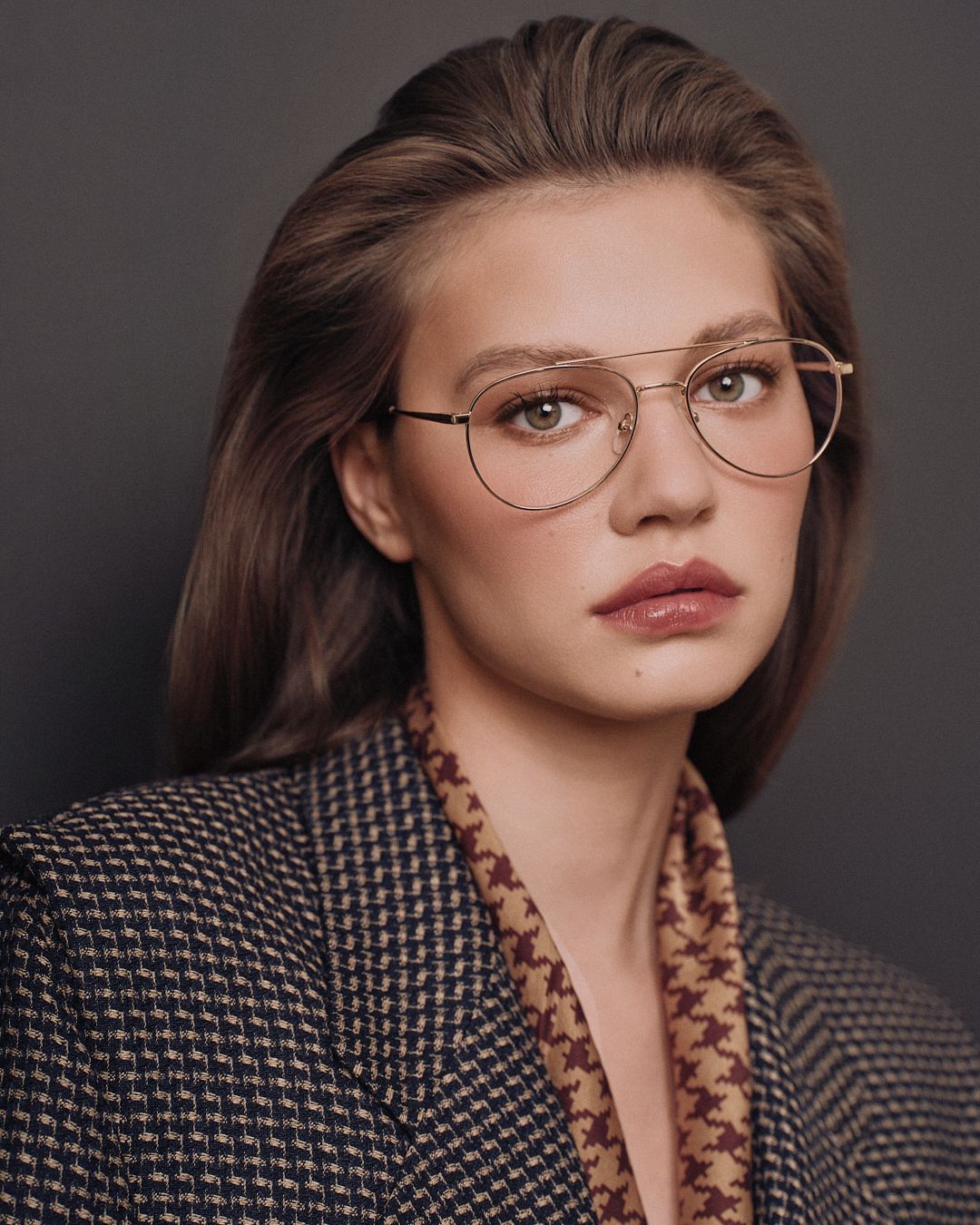 Young woman with brown hair wearing gold aviator glasses and a houndstooth blazer poses against a gray background.