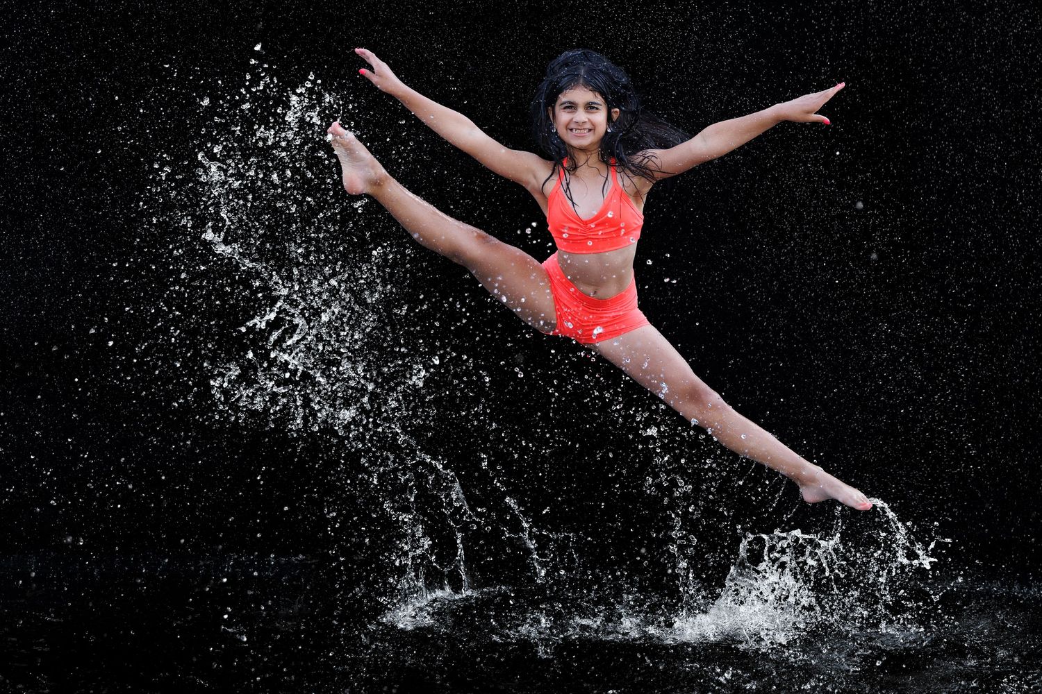 Young female dancer leaps mid-air in orange bikini, surrounded by dramatic water splashes against black background.