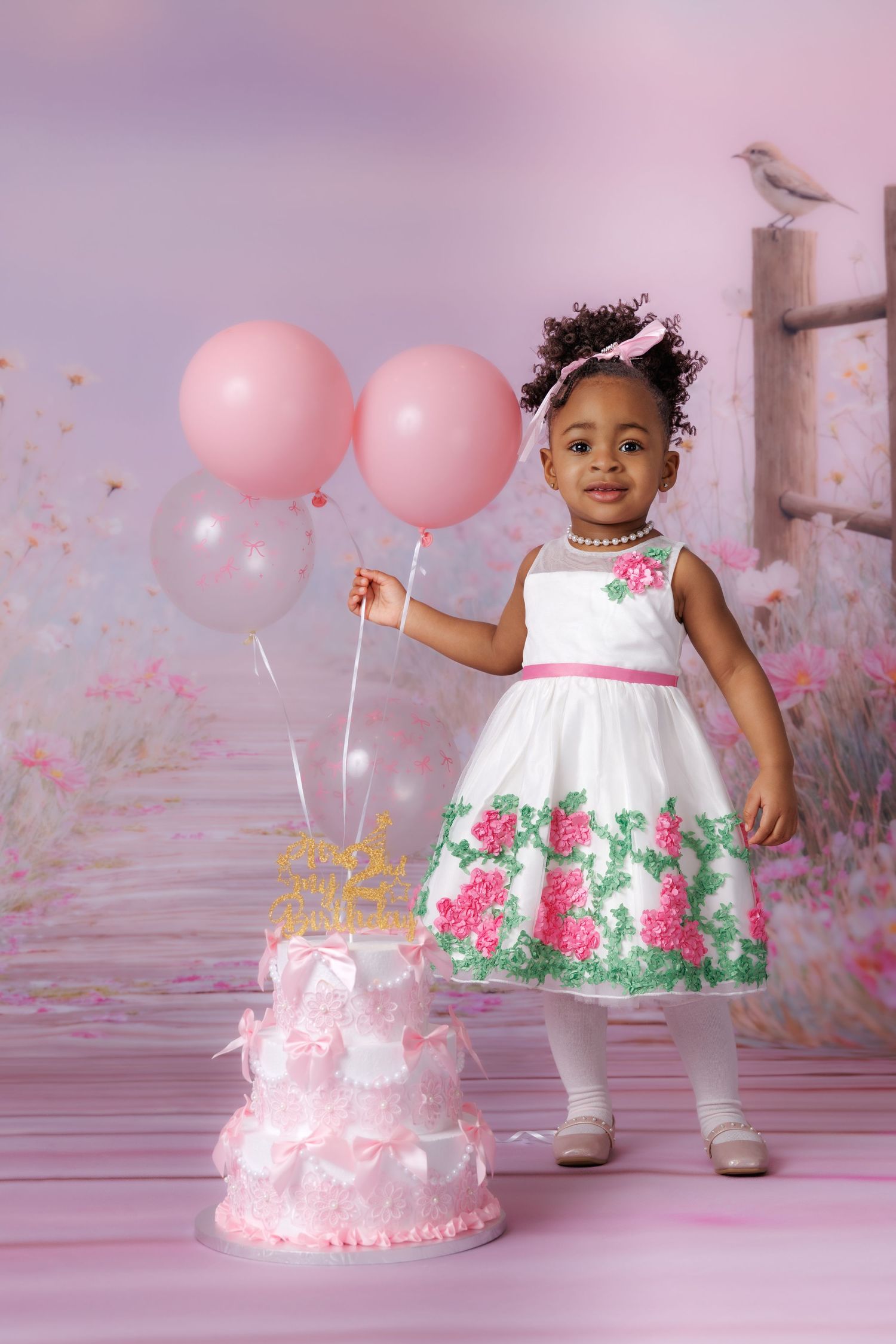 Toddler girl in white floral dress holds pink balloons beside a tiered birthday cake in a dreamy pink studio setting.