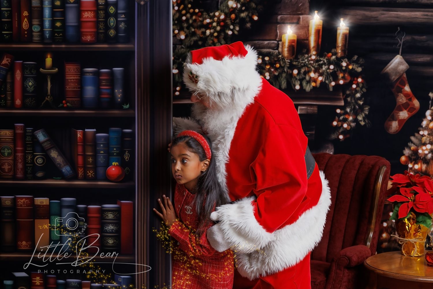A young girl in a red dress poses with Santa Claus near a bookshelf and decorated Christmas tree in a cozy holiday setting.