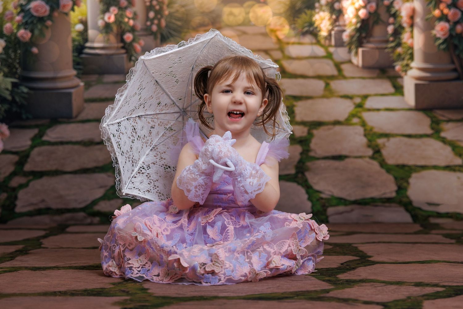 Happy toddler girl in a purple dress holding a white lace umbrella sitting on a garden stone pathway.