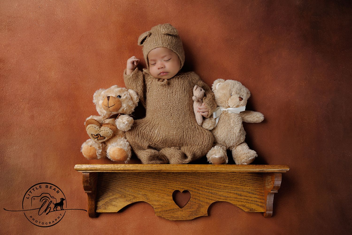 Adorable sleeping newborn in brown knit outfit posed on wooden shelf surrounded by teddy bears.