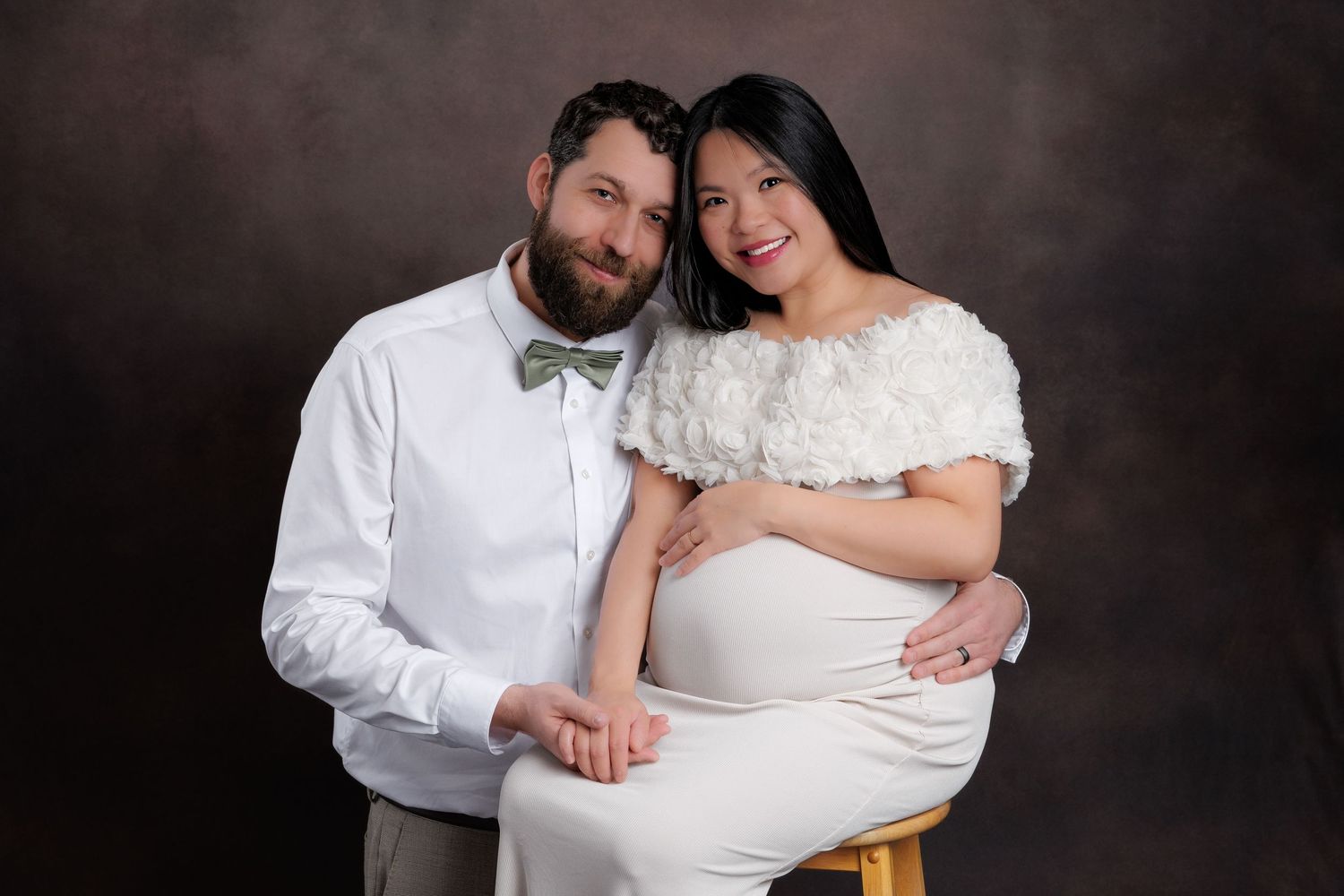 Expecting couple posing together, man in white shirt with bow tie and woman in white maternity gown.