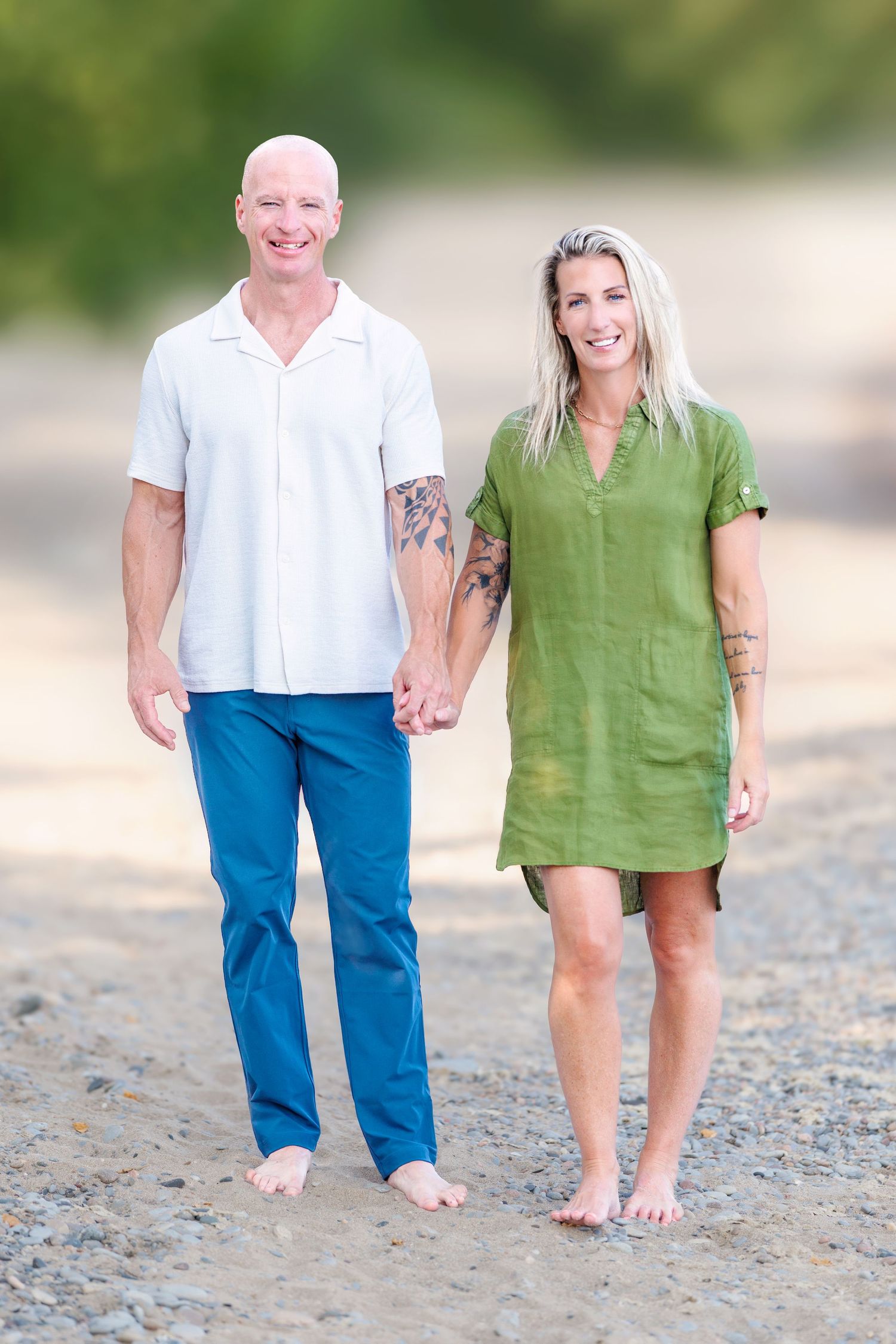 Bald tattooed man holds hands with blonde woman in green dress while walking barefoot on a beach.