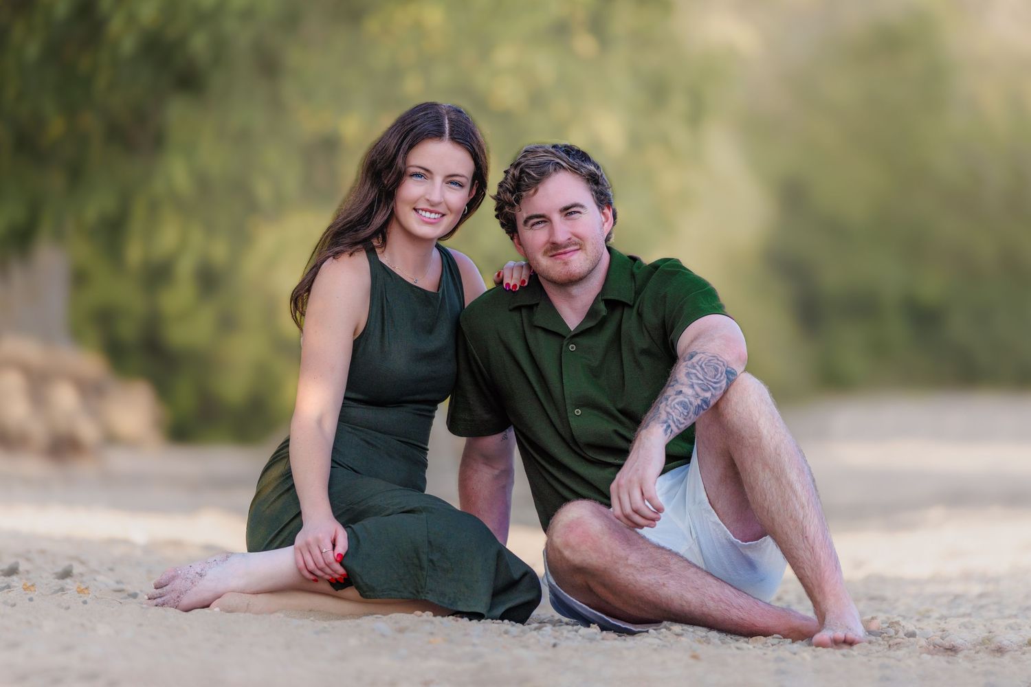 Smiling young couple sitting on sandy beach, woman in green dress and man in green polo shirt.