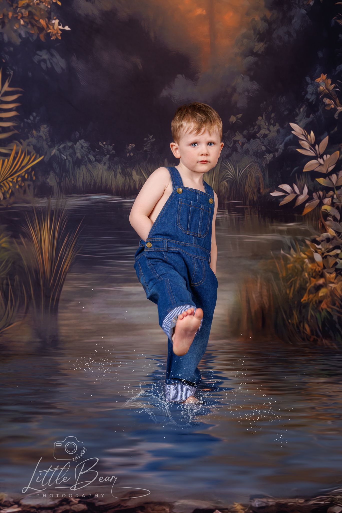 Toddler boy in denim overalls splashing barefoot in a scenic woodland stream with autumn foliage backdrop.
