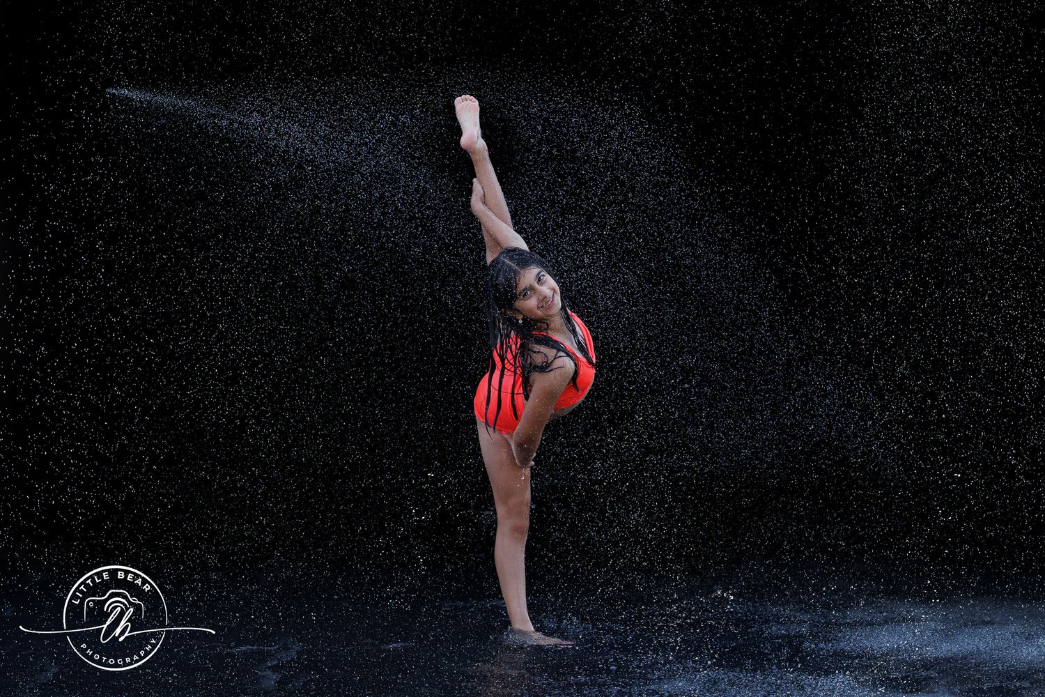 Gymnast in red leotard performs elegant leg hold pose in dramatic rain shower against dark background.