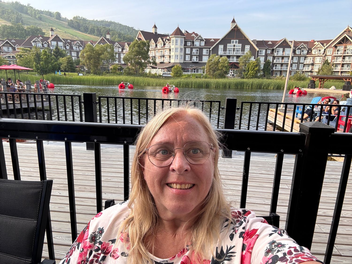 Smiling woman with glasses takes a selfie on a waterfront deck with European-style resort buildings and red kayaks behind her.