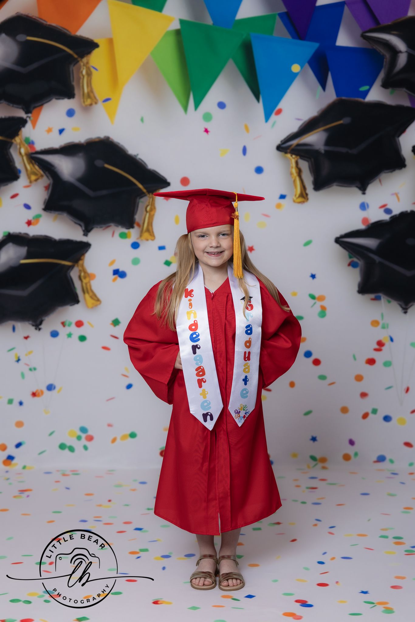 Young girl in red kindergarten graduation cap and gown poses with colorful confetti and mortarboard balloons.
