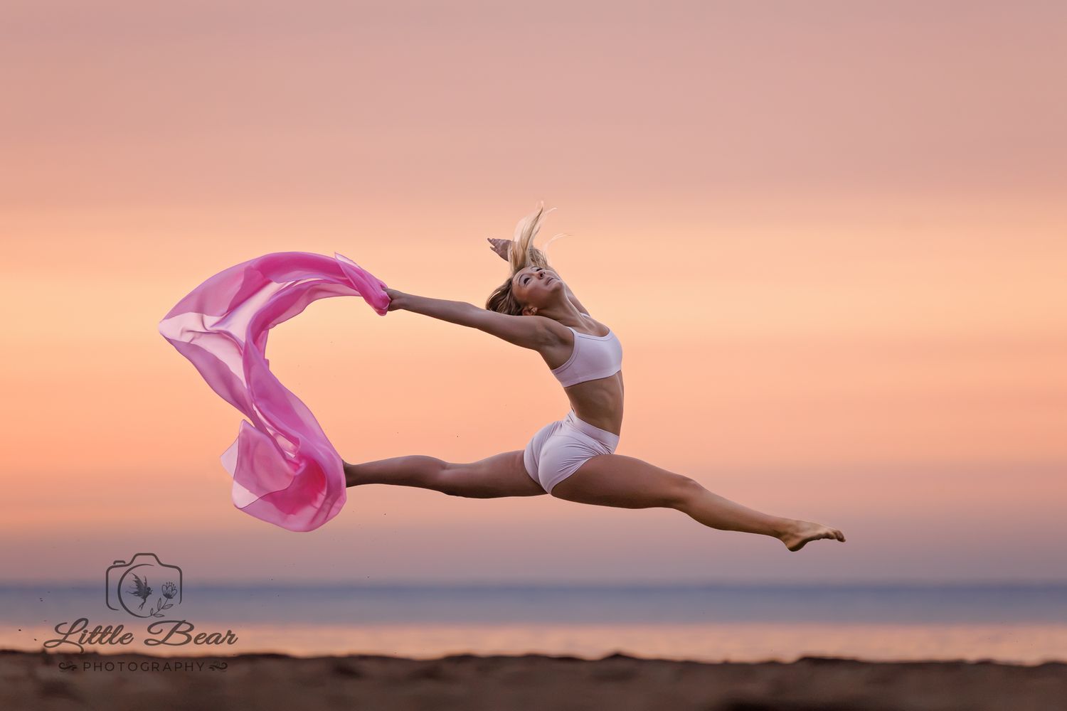 Graceful dancer leaps at sunset on beach, holding flowing pink fabric, wearing white outfit, captured by Little Bear Photography.