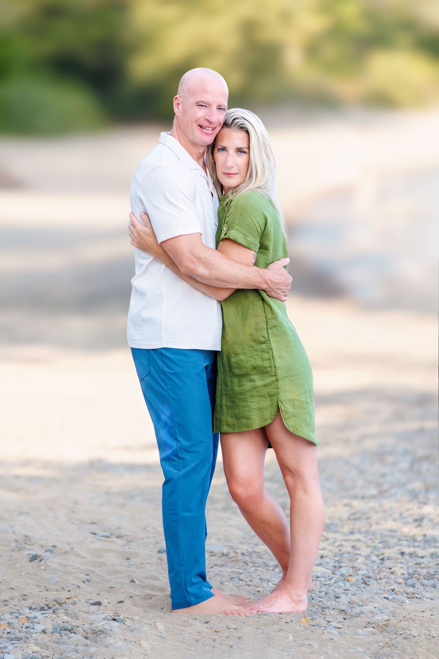 Bald man in white shirt embraces blonde woman in green dress while posing on a sandy beach.