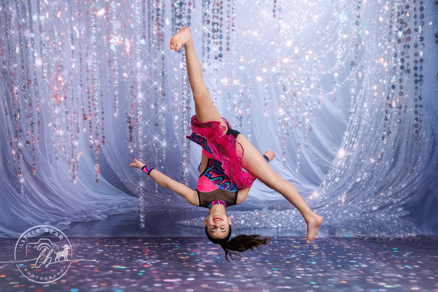 Dancer in colorful costume performs inverted aerial pose on glittery backdrop.