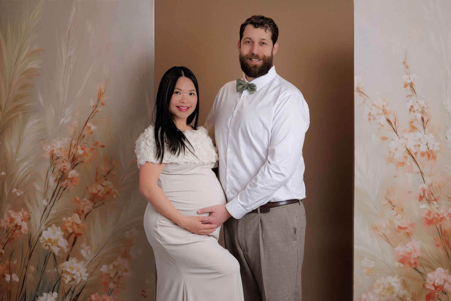 Expecting couple in elegant attire posing together in front of a floral and neutral-toned maternity backdrop.