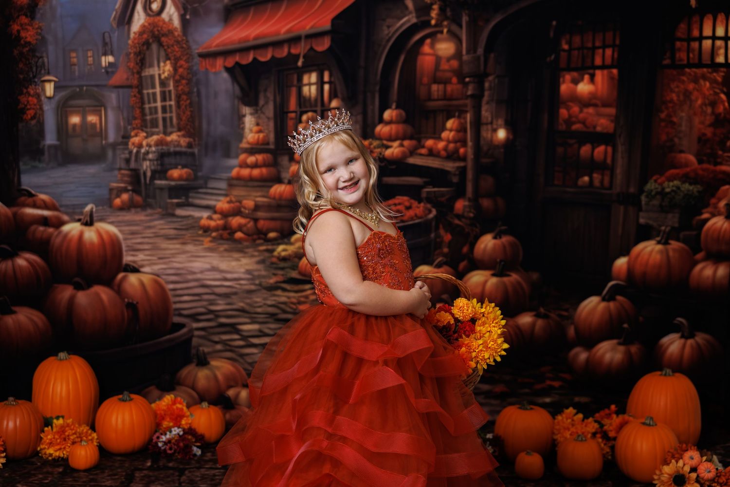 Girl in red dress smiles among pumpkins in a festive Halloween-themed photo with a spooky village backdrop.