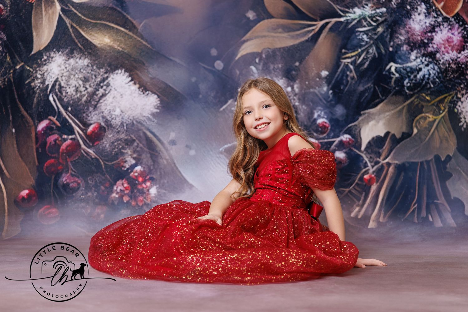 A cheerful girl in a sparkling red gown sits gracefully against a dreamy winter floral photography backdrop.