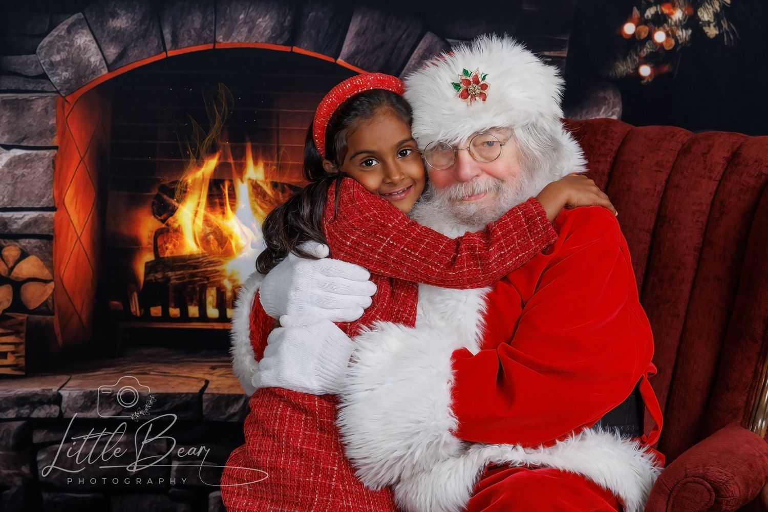 A young girl hugs Santa Claus beside a warm glowing fireplace in a cozy Christmas holiday portrait photo.