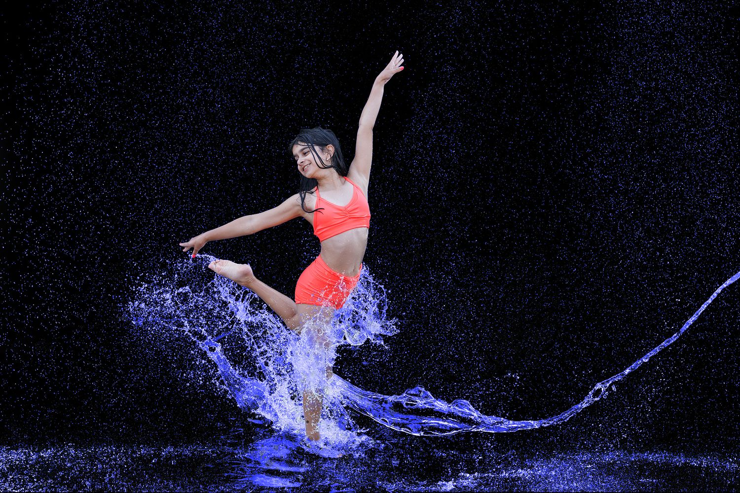 A dancer in orange activewear leaps gracefully through water splashes against a dark background.