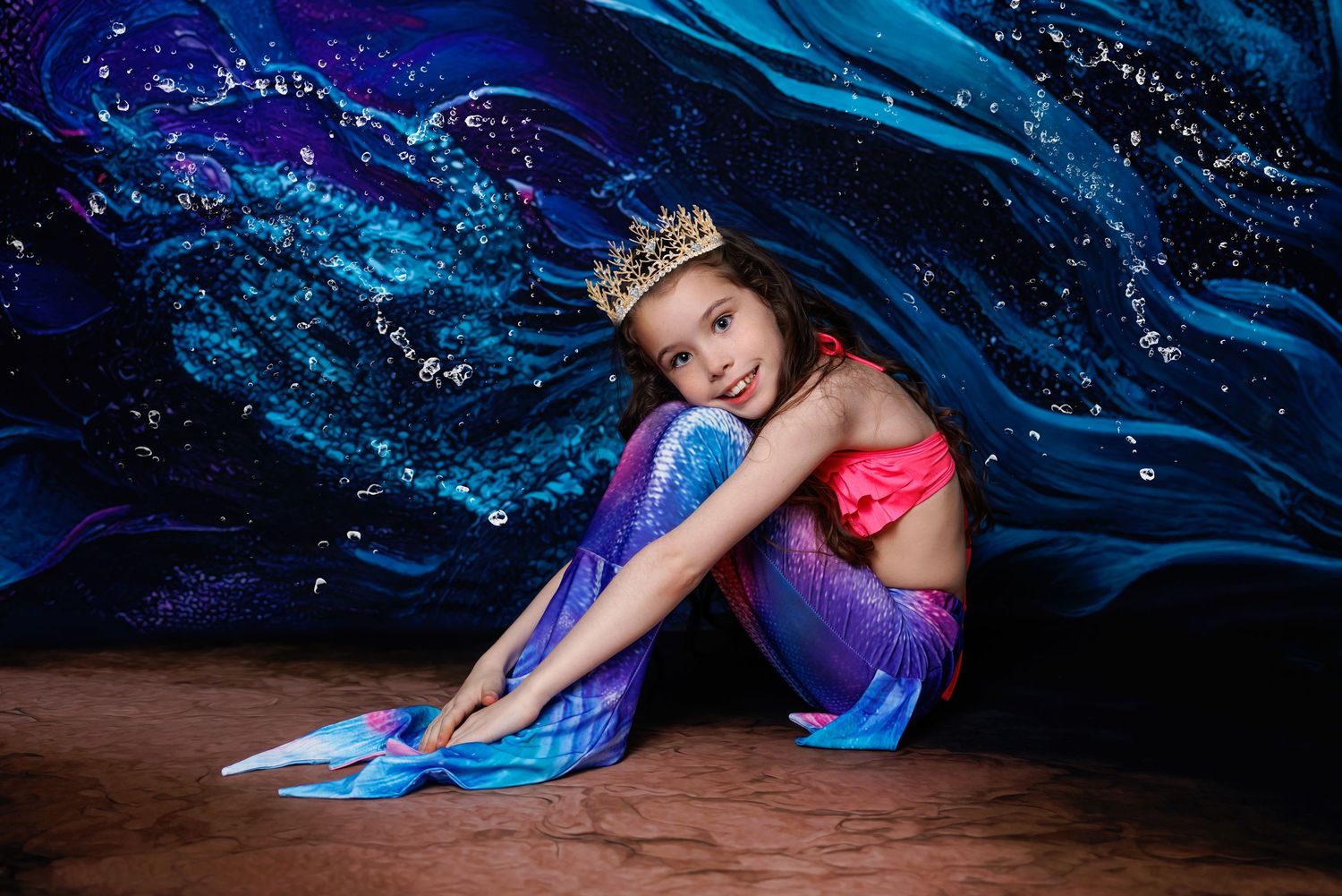 Girl in mermaid tail and gold crown sits on floor against stunning blue and purple ocean backdrop.
