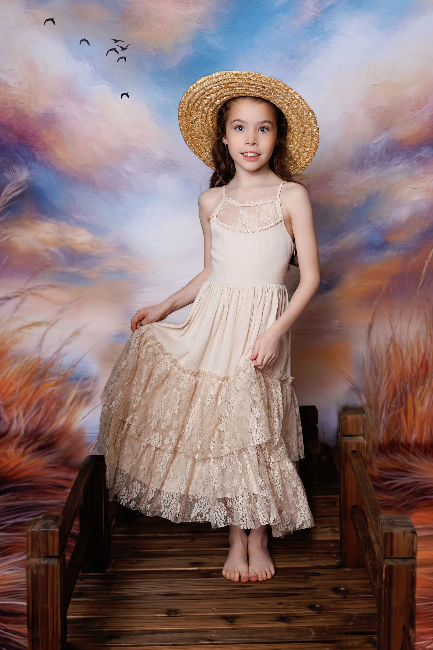 A young girl in a cream lace dress and straw hat poses barefoot on a wooden bridge against a colorful sky backdrop.