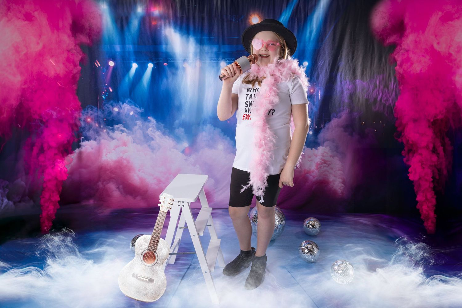 Child performer in black hat and Taylor Swift shirt holds pink feather boa and microphone on colorful smoky stage.