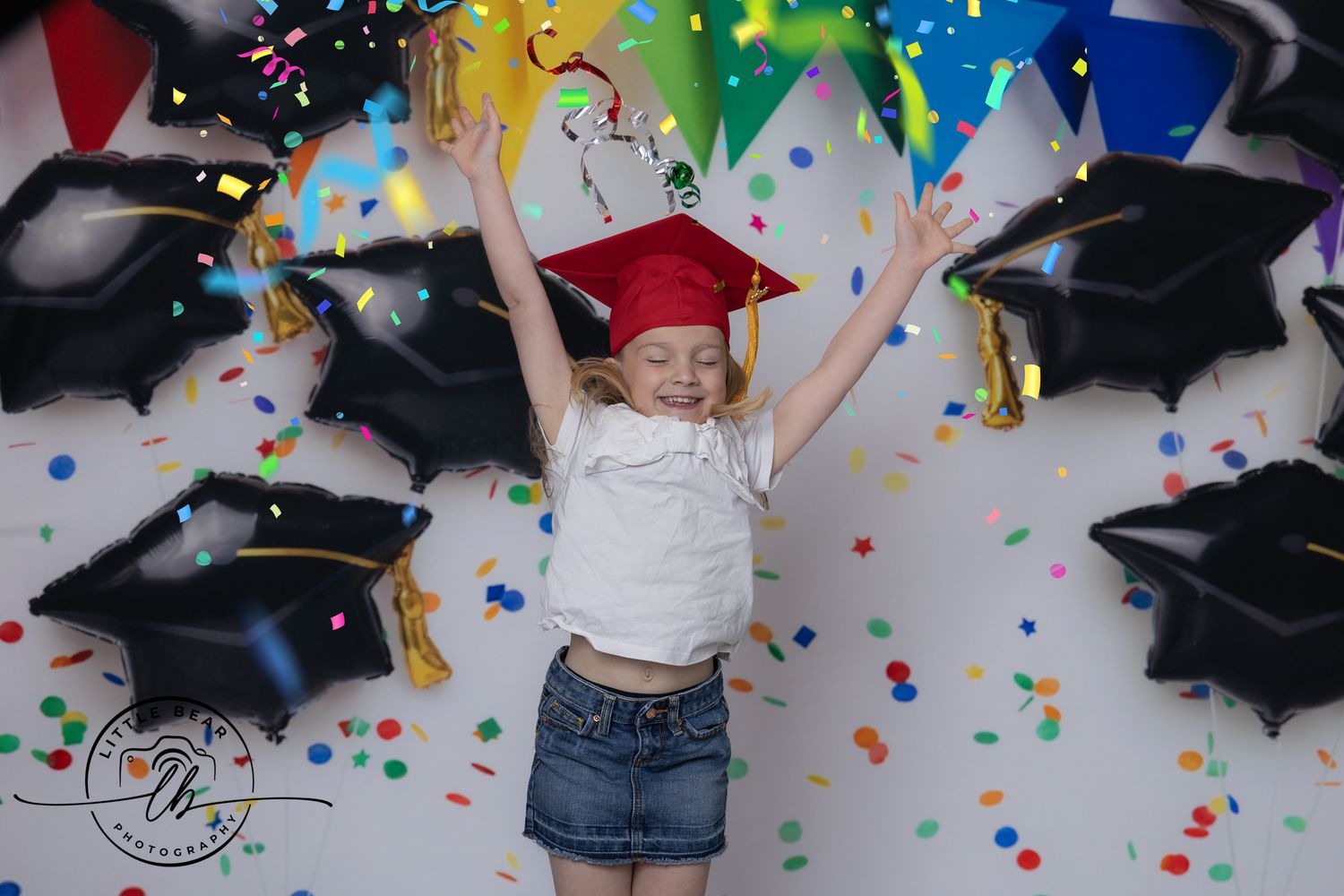 Young girl in red graduation cap jumps joyfully amid colorful confetti and black mortarboard balloons.