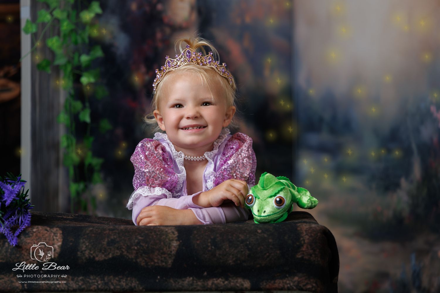 Toddler girl in Rapunzel princess costume with crown holds green frog toy, smiling at camera in enchanted forest setting.