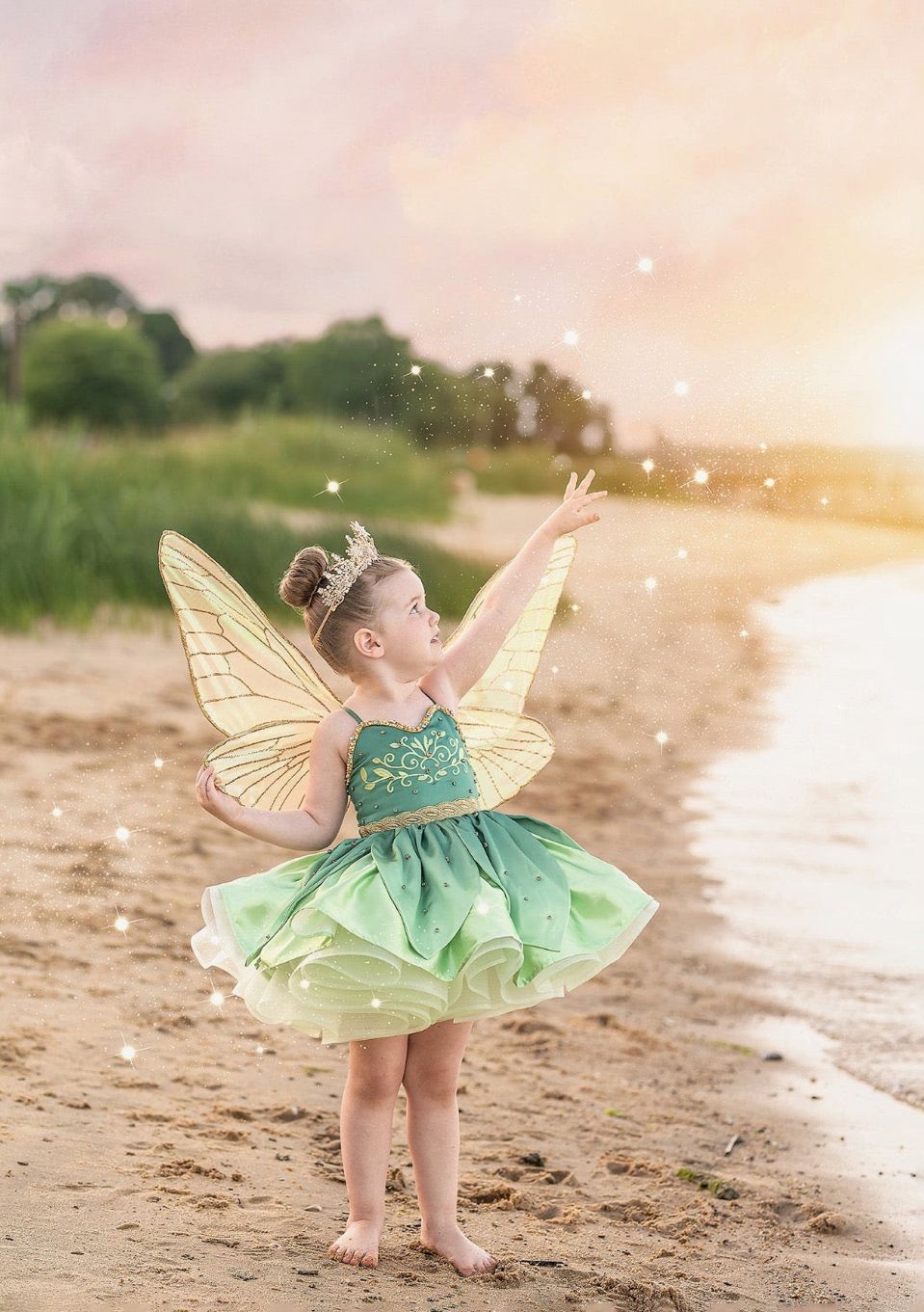 A young girl in a green fairy costume with yellow wings stands on a beach at sunset, reaching toward sparkling magic dust.