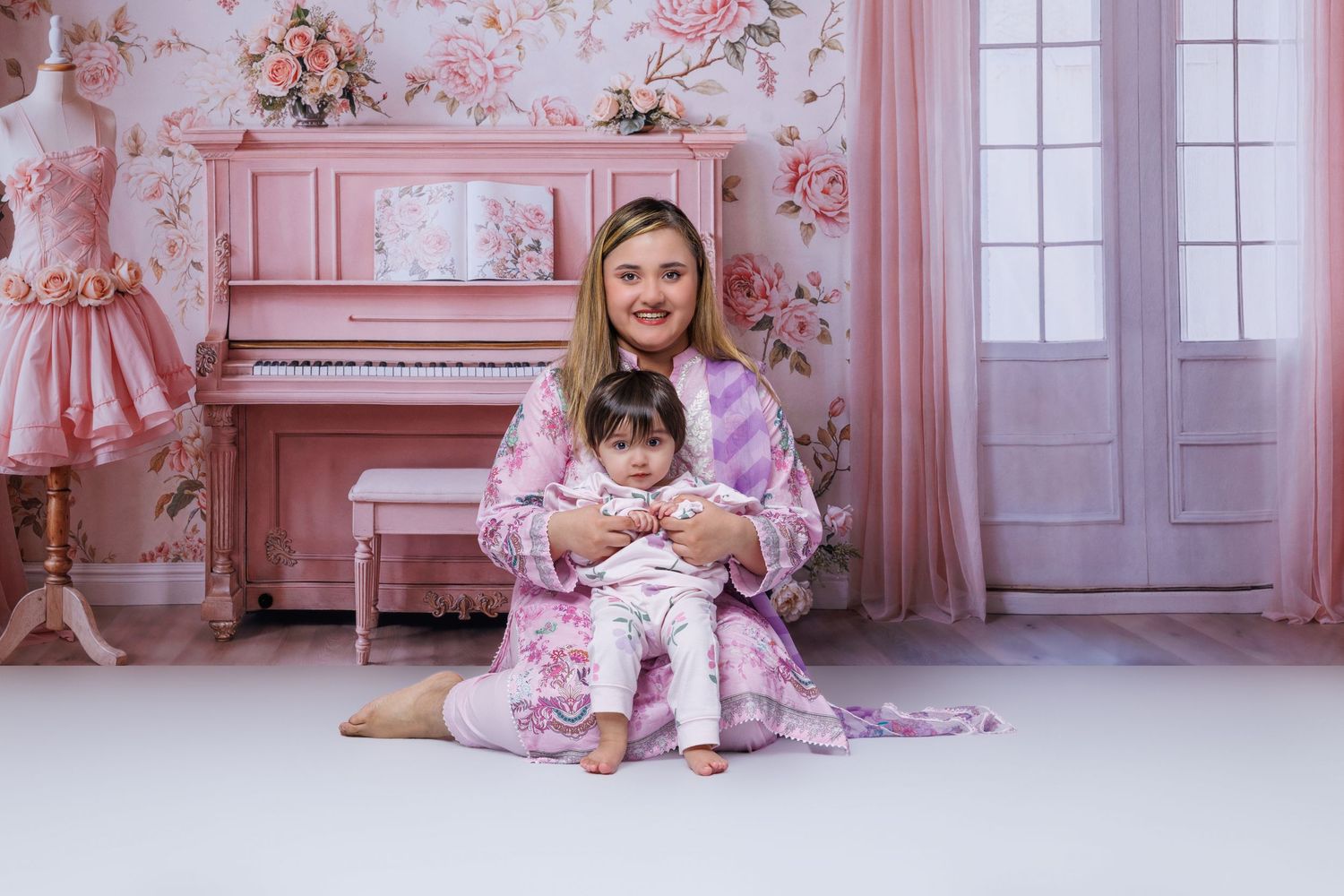 A smiling woman holds a baby girl on her lap in a pink floral decorated photography studio.