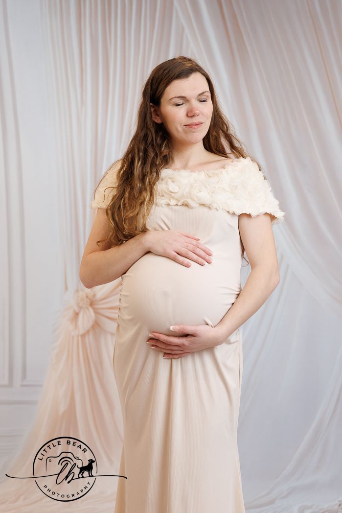 Pregnant woman in elegant cream off-shoulder gown cradling her baby bump in a professional maternity photo session.