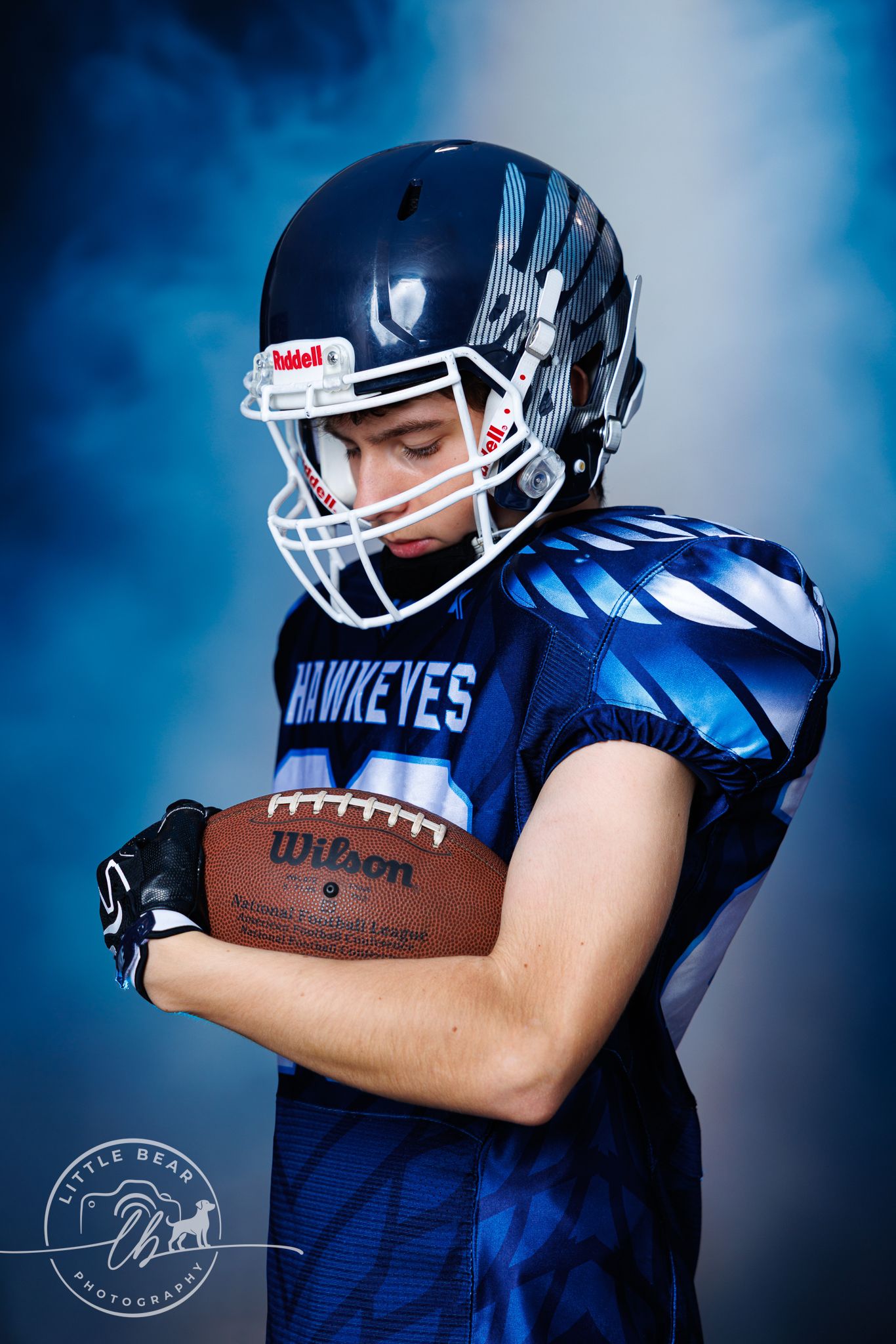 Youth football player in blue Hawkeyes uniform holding Wilson NFL football while looking down dramatically.
