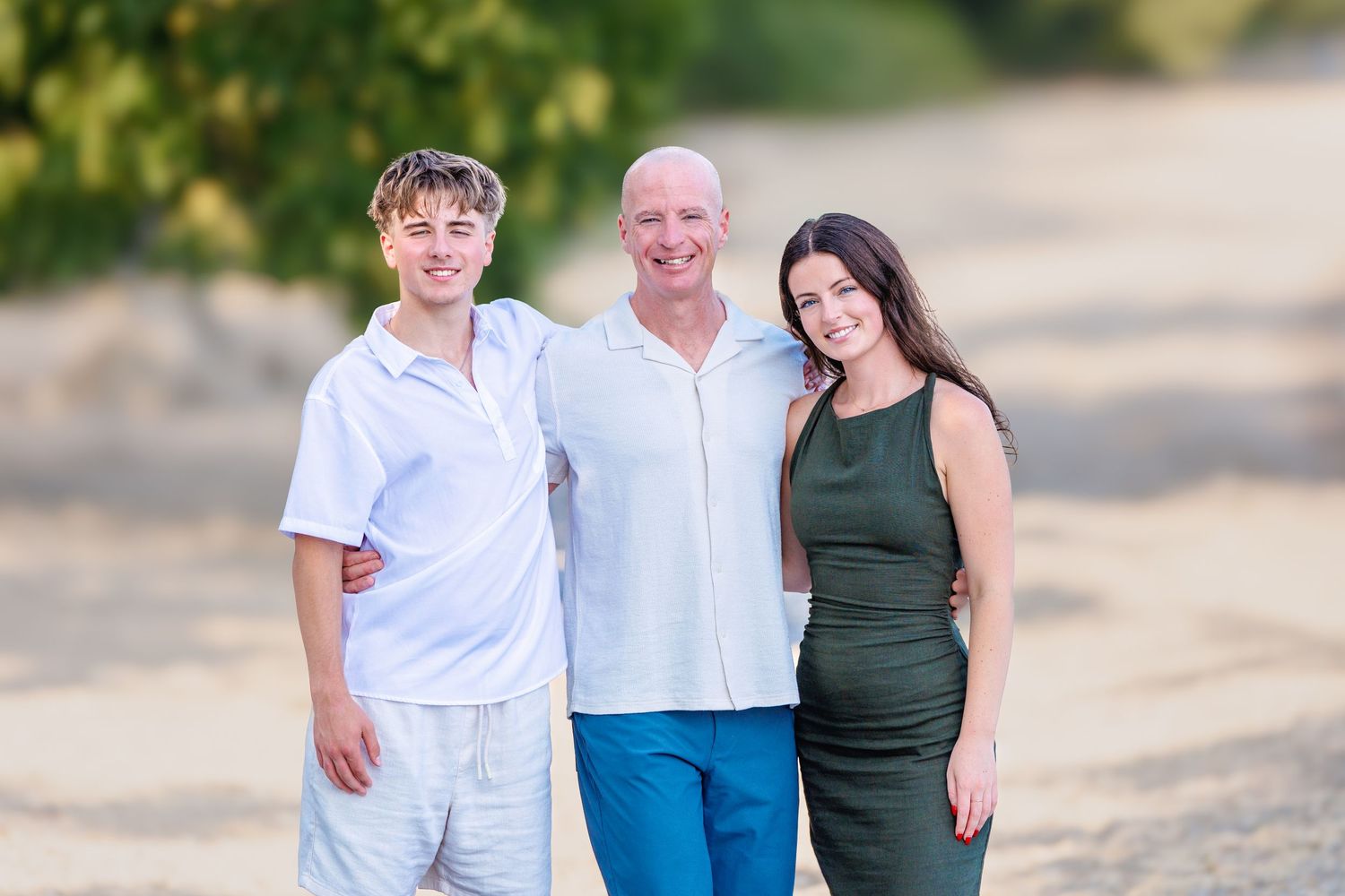 A man poses on a sandy beach with a teenage boy and girl smiling together.