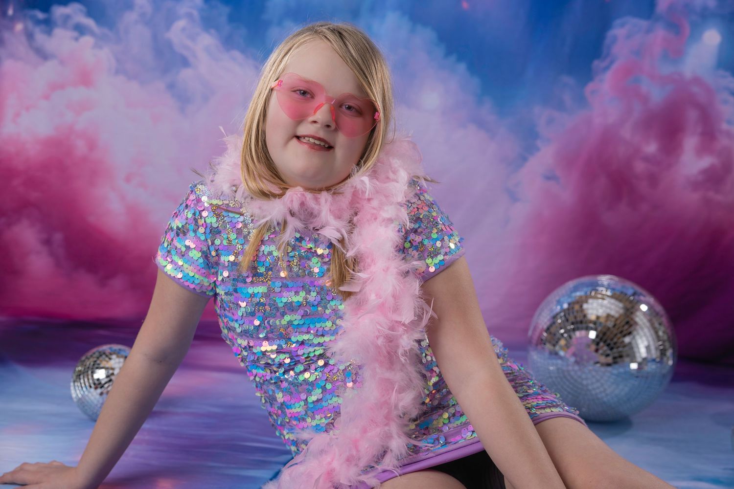 Smiling blonde girl in sequin dress and pink heart sunglasses poses with feather boa and disco balls.