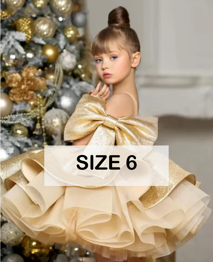 Girl in size 6 gold ruffled ball gown dress with bow, posing near Christmas tree with ornaments.