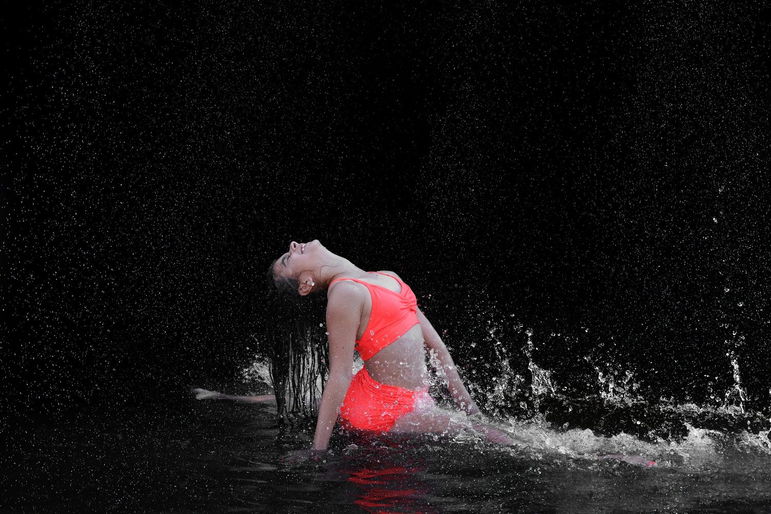 A dancer in a red bikini emerges from water with dramatic splashes against a dark background.