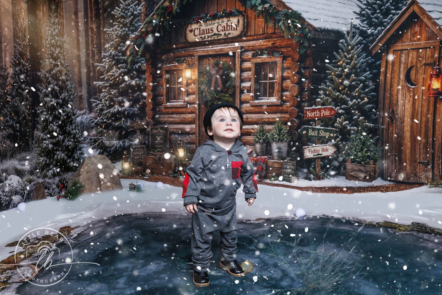 Toddler boy in gray outfit stands in snowy Christmas village scene with rustic log cabin and festive decorations.