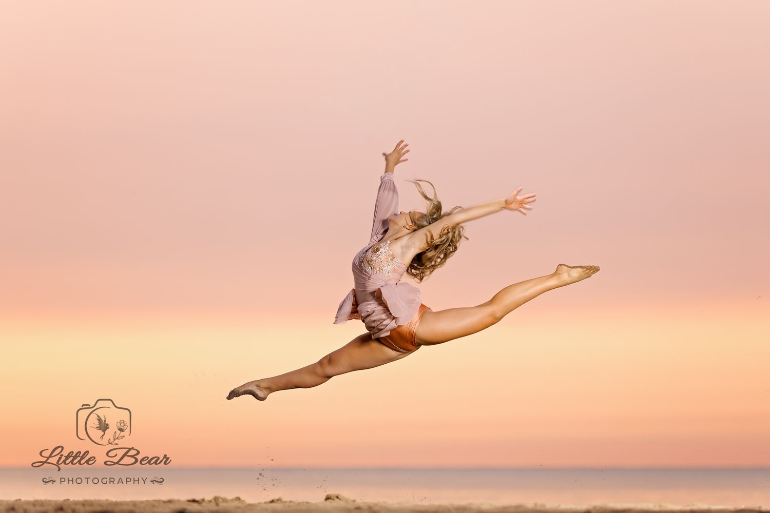 A dancer leaps gracefully against a pink sunset sky over the beach, performing a stunning split jump.