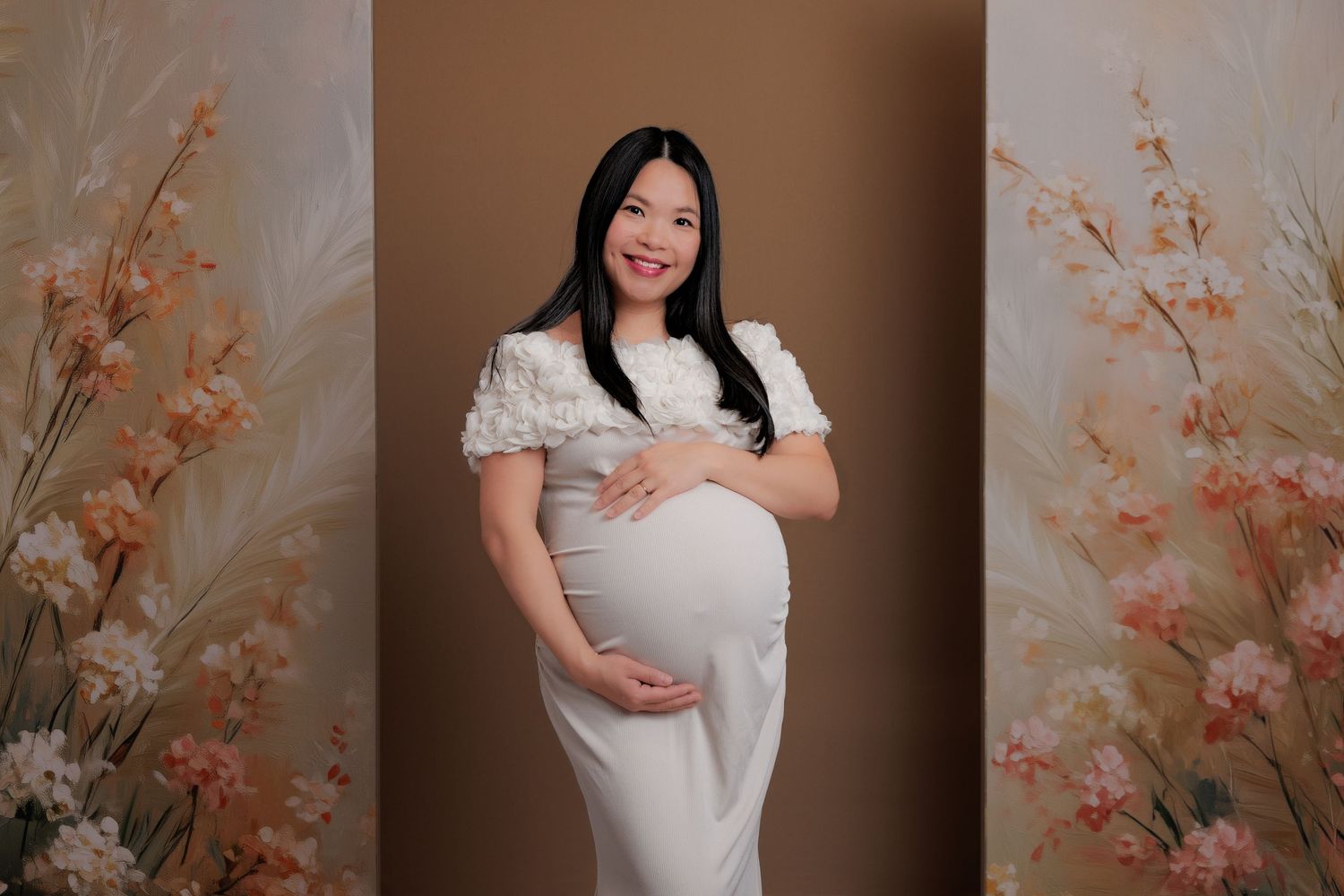 Smiling pregnant woman in white maternity dress posing between floral panels in professional studio photo.