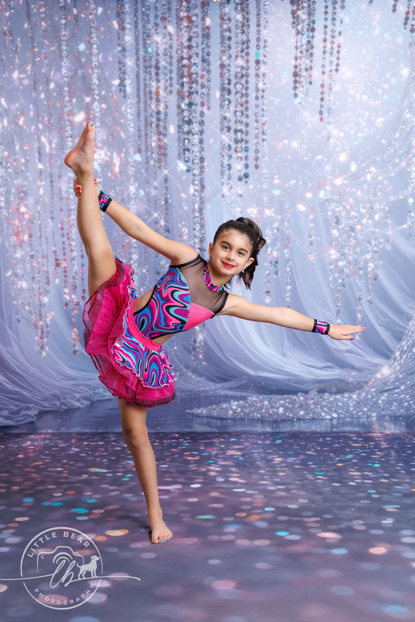 Young dancer in pink and blue costume performs high leg hold on glittery stage.
