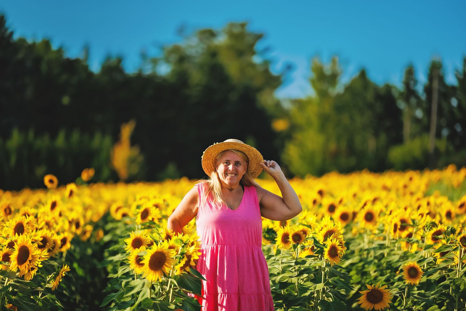 Woman in pink dress and straw hat smiling in a vibrant sunflower field on a sunny day.