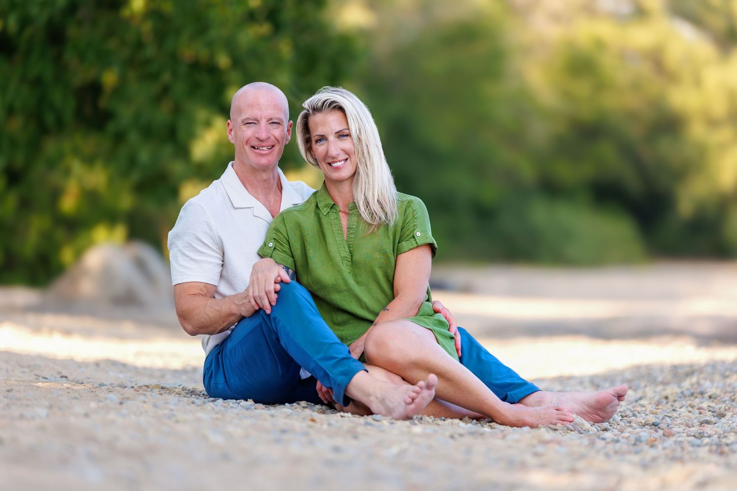 Smiling couple sitting together on sandy beach, man in white shirt and woman in green outfit.
