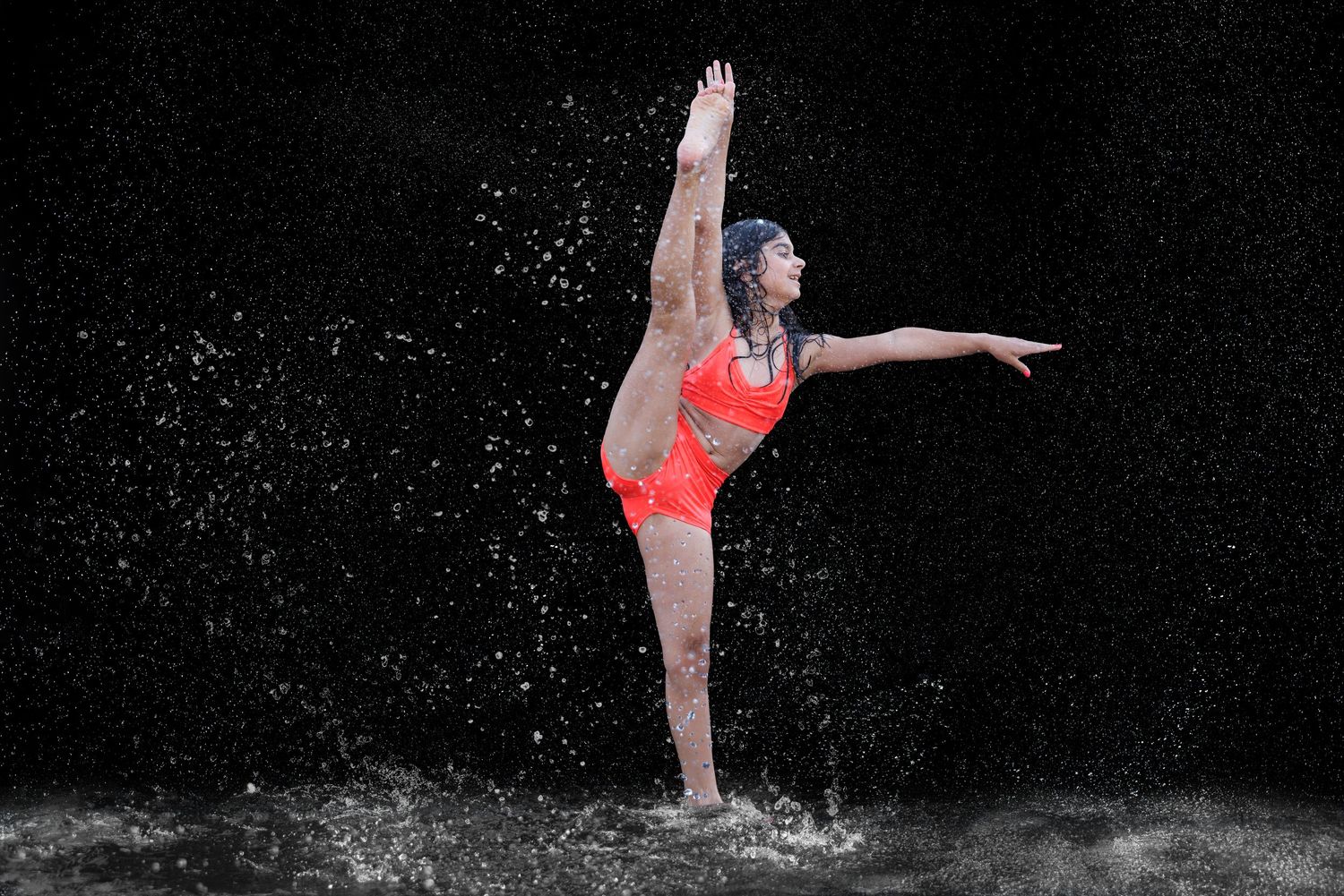 Synchronized swimmer in red swimsuit performs elegant split leap with water splashing against black background.