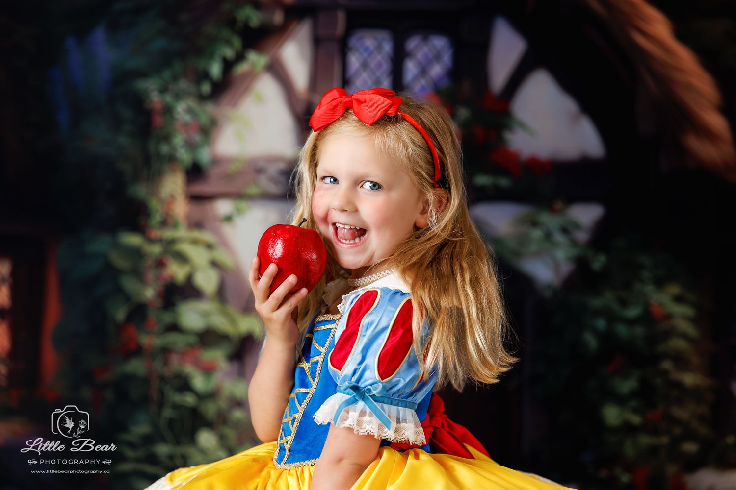 Girl in Snow White costume holds red apple, smiling, with storybook backdrop, captured by Little Bear Photography.