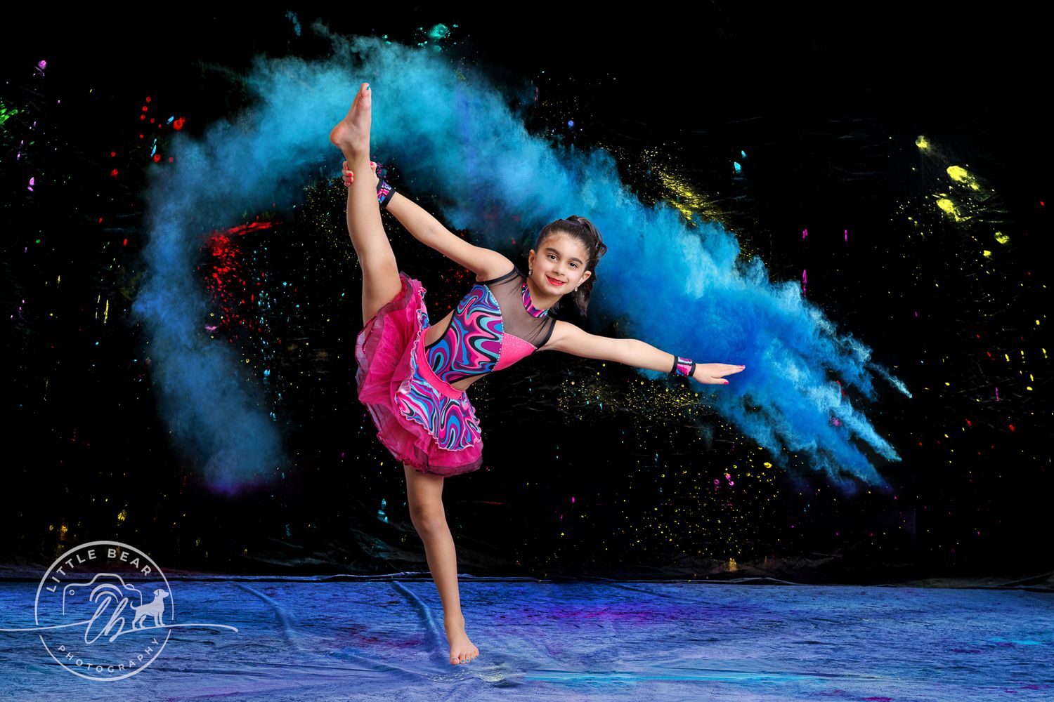 Young dancer in pink costume performs leg hold amid colorful powder explosion.