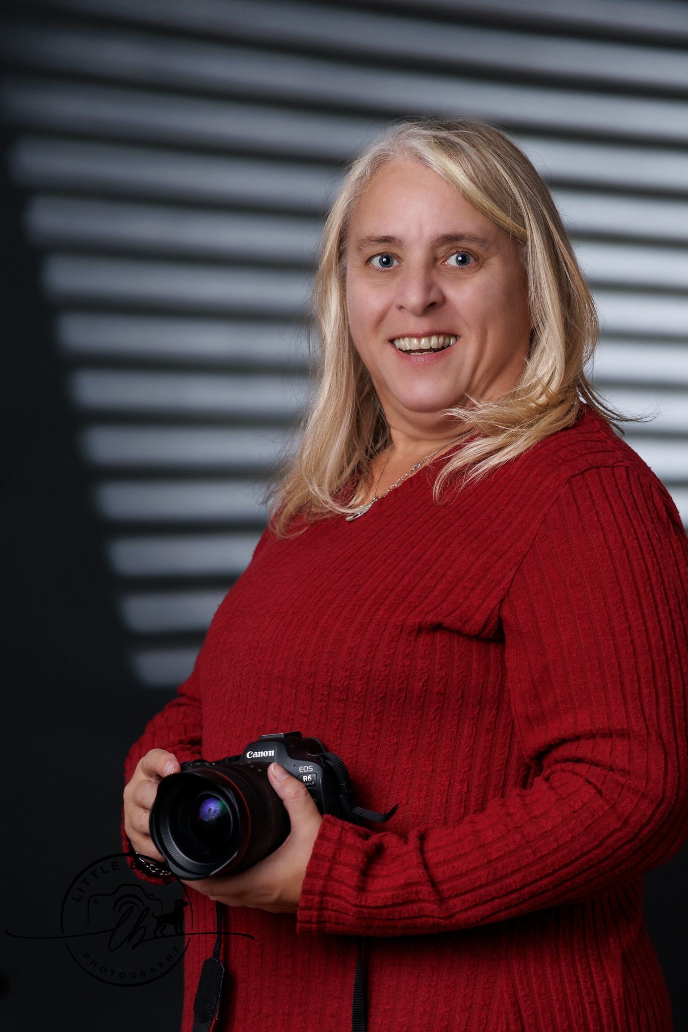 Smiling blonde woman photographer holding Canon camera wearing red sweater in studio.