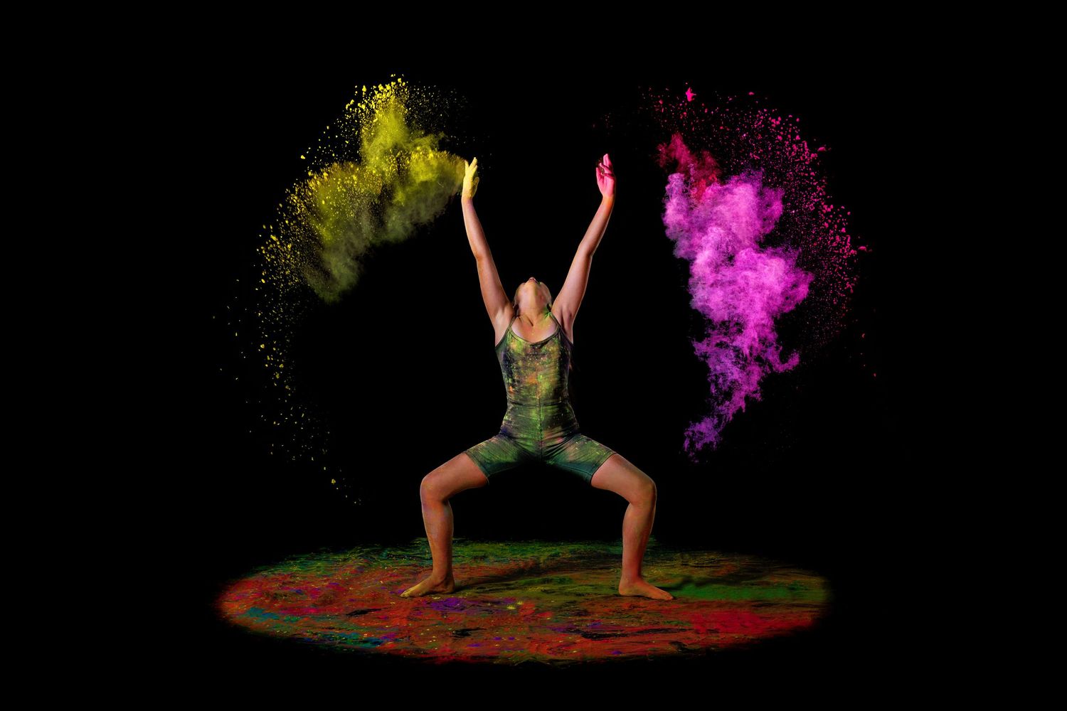 Dynamic dancer in colorful outfit strikes powerful pose with yellow and pink powder explosions on dark background.