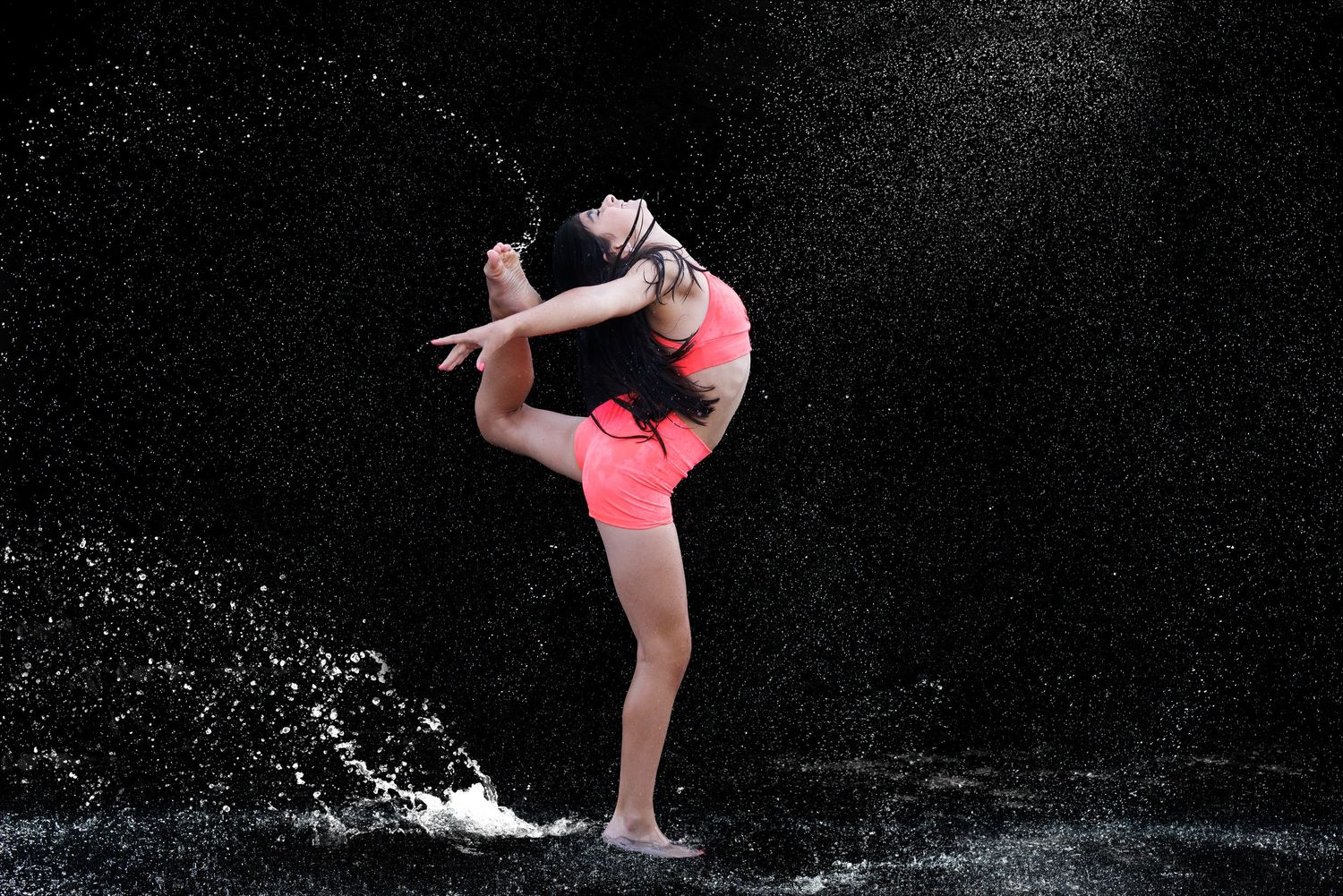A gymnast in pink athletic wear performs a graceful back bend while water splashes around her on a dark background.
