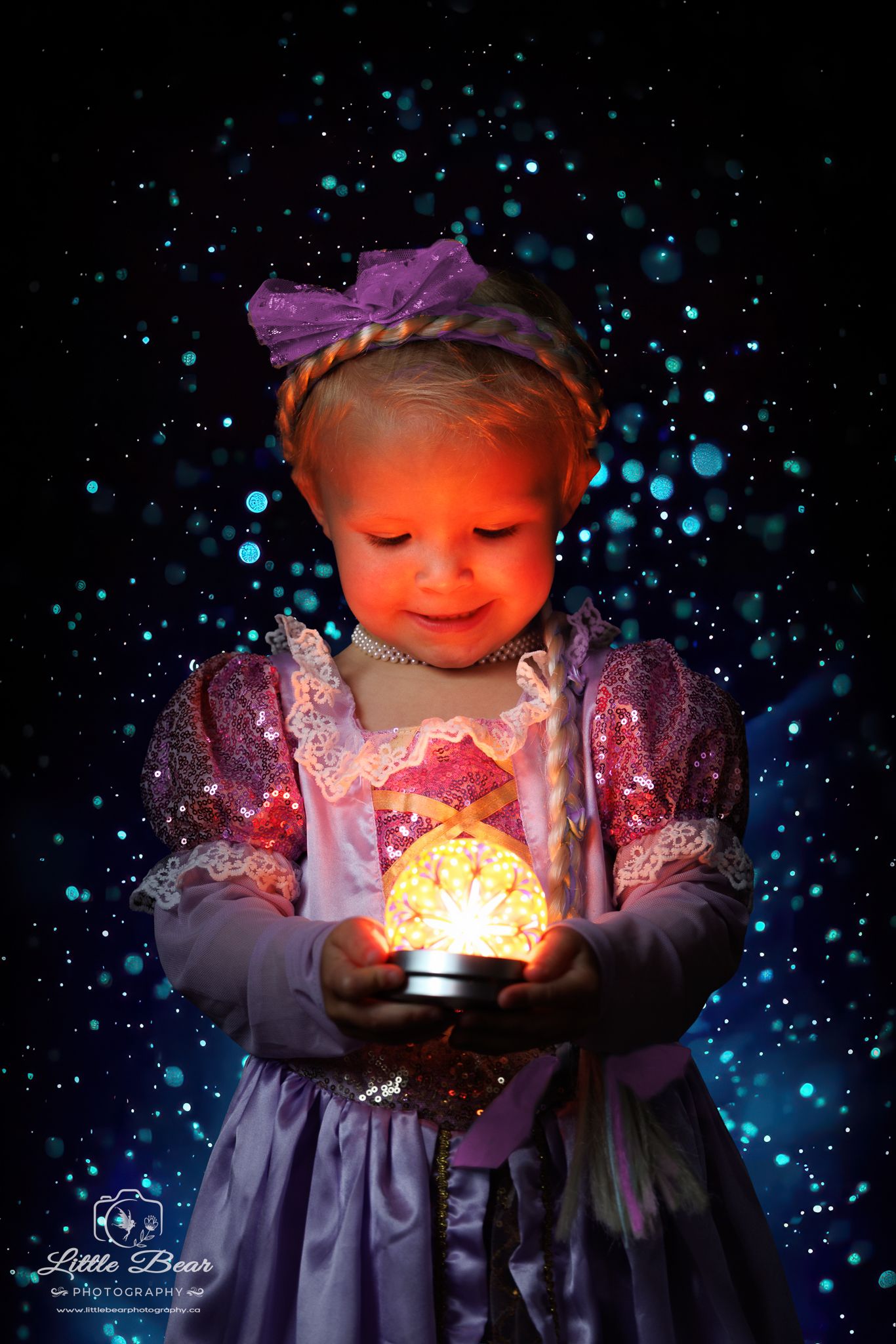 A toddler girl in a Rapunzel costume holds a glowing orb, illuminated by magical bokeh lights on a dark background.